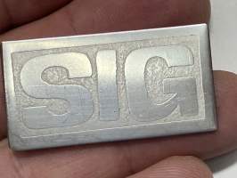 SIG-rintamerkki, Matti &quot;Inksu&quot; Inkisen (Inkinen) perustaman yhtyeen ensimmäisiä &quot;fanituotteita&quot;, metallinen rintamerkki, tekijä Seppo Määttänen (&quot;Raision Rembrandt&quot;)